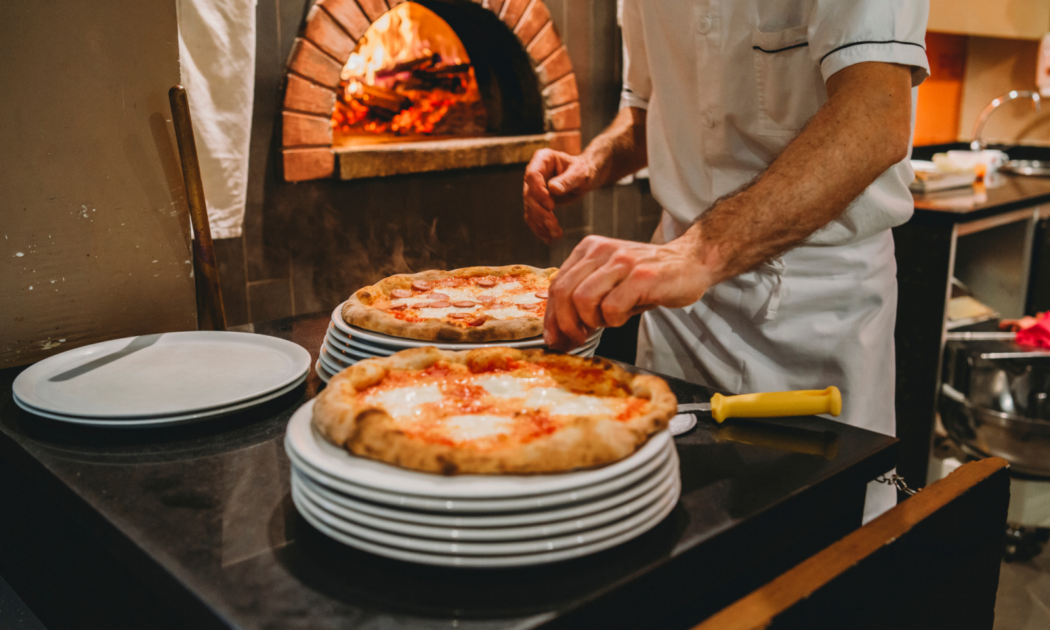Photo of a pizza chef preparing two pizzas to put in the oven behind.
