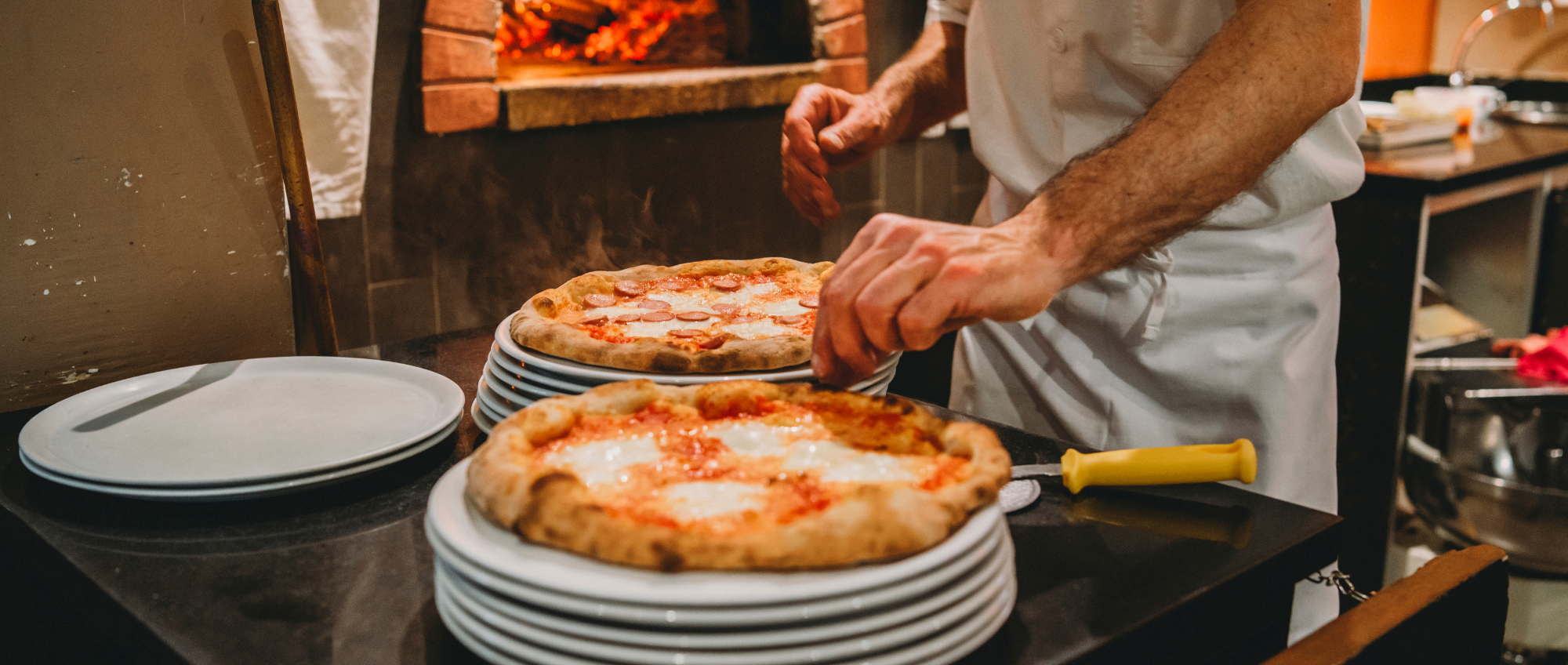 Photo of a pizza chef preparing two pizzas to put in the oven behind.