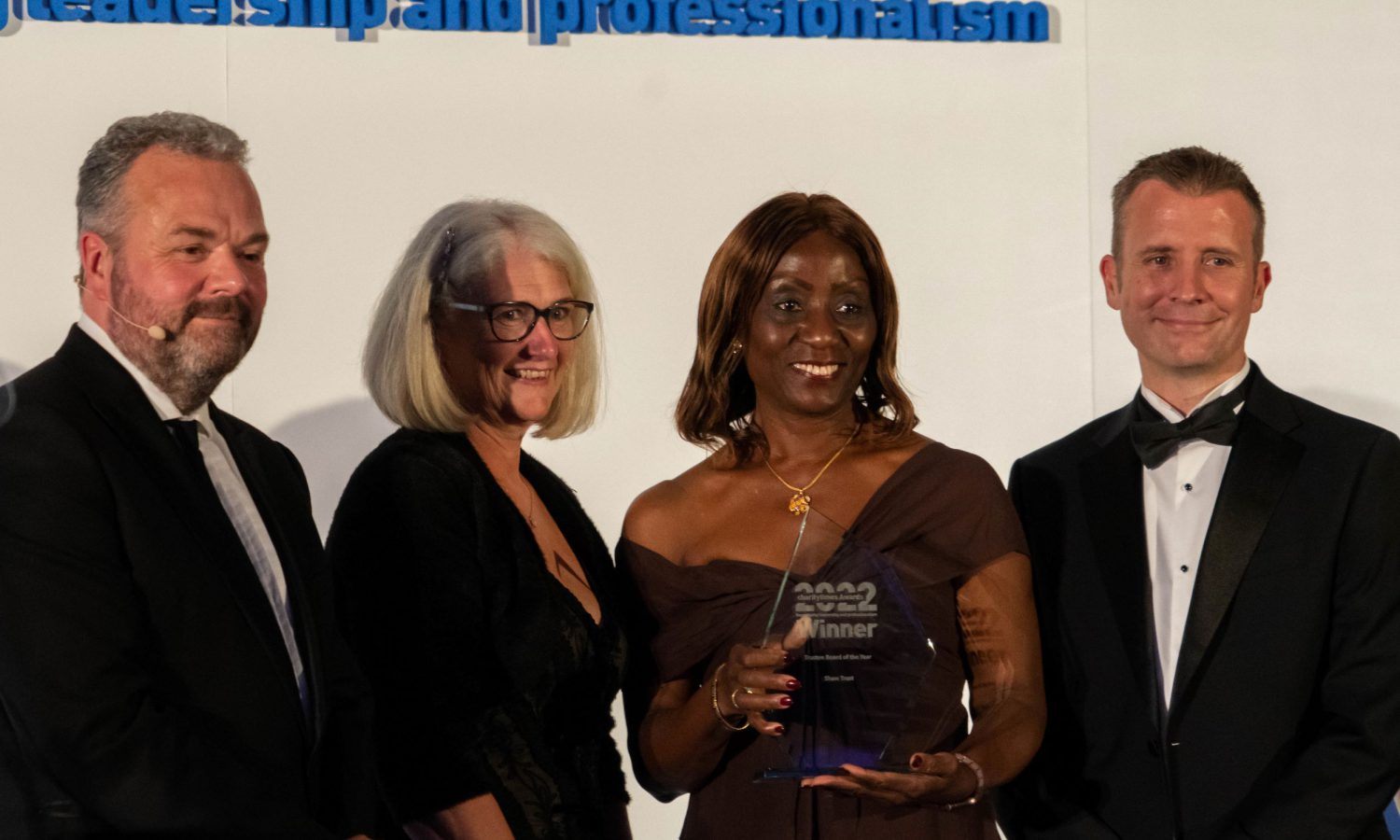 Image of four people at Awards ceremony. They are looking sideways from the camera. They are dressed in smart black tie, and are smiling. One woman is holding an award