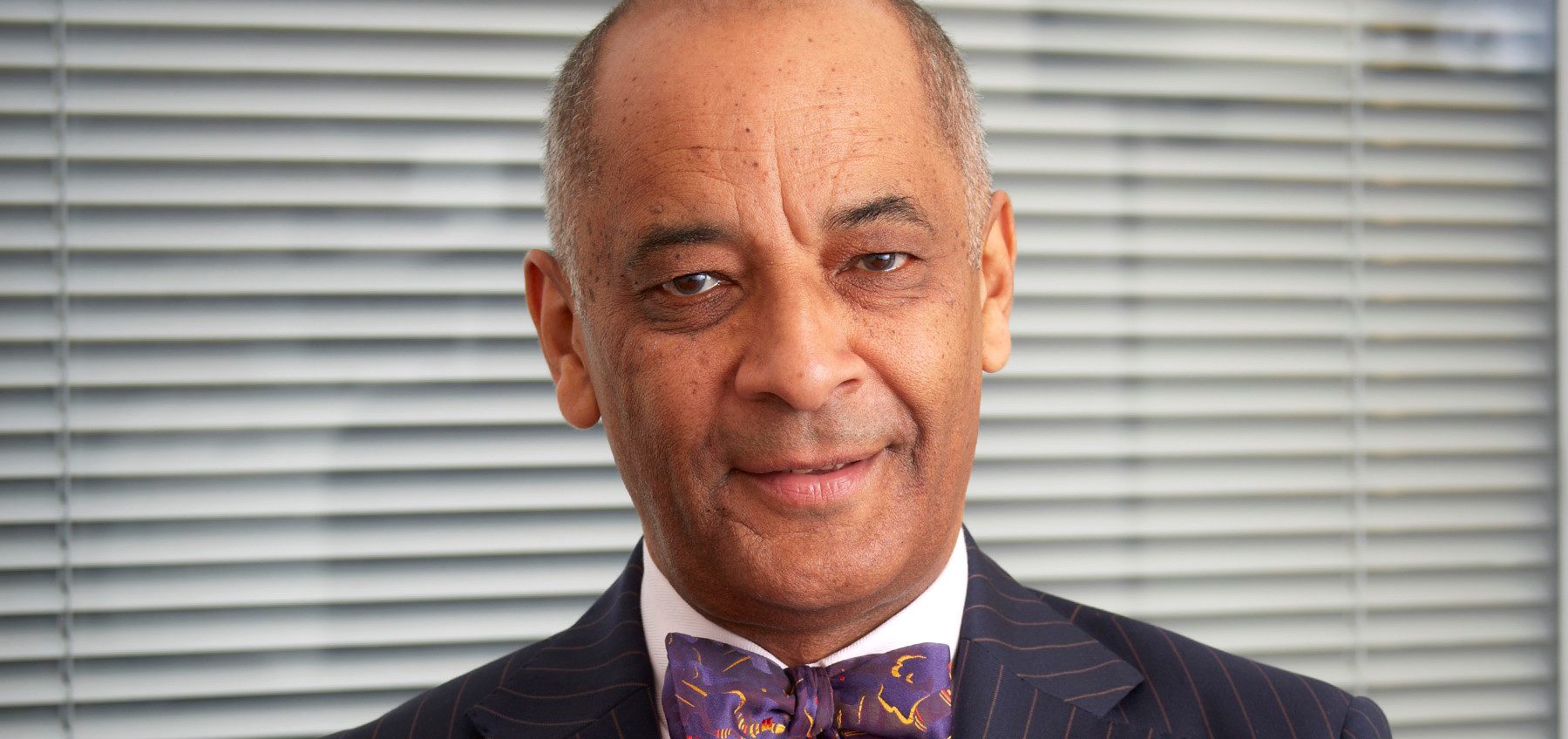A smiling Sir Ken Olisa is dressed in smart clothes with a purple bow tie.