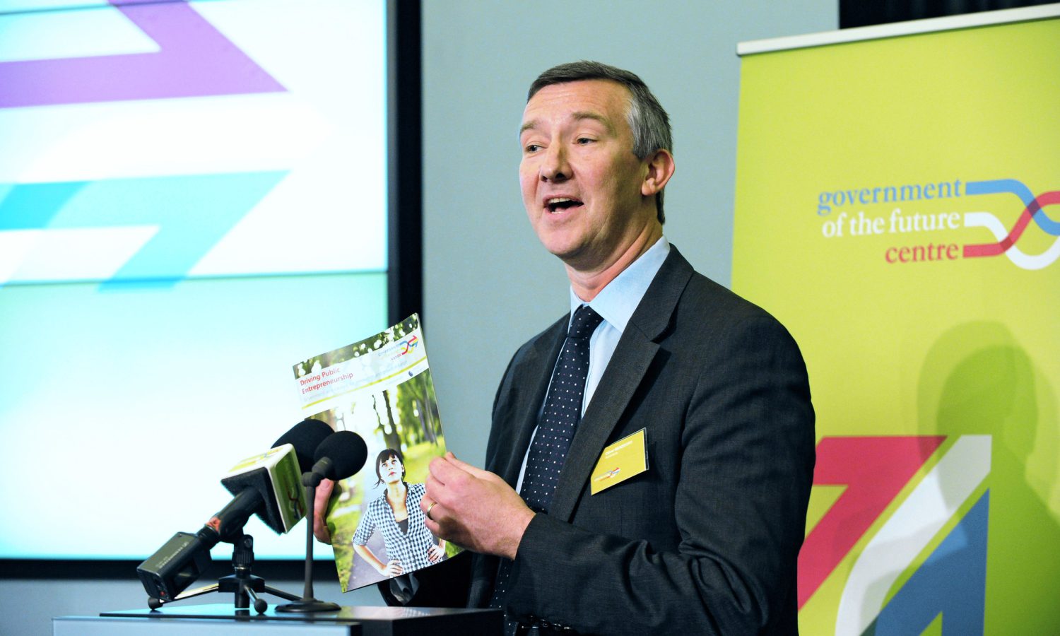 Man in smart suit stands at lecturn and talks into a microphone