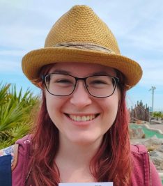A photo of a lady with red hair down over her shoulders. She is wearing a brown sun hat and glasses and smiling straight to the camera.