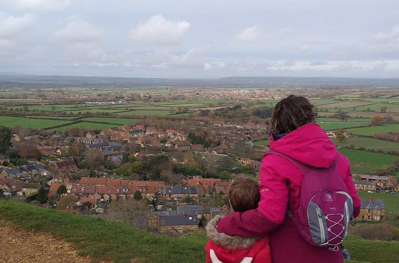 A woman in a pink coat with a small pink backpack on her back. Her left arm is held around a small boy in a red, white and navy colour block coat. They are looking out over the countryside. The sky is grey with low white clouds and small villages are dotted in amongst green fields.