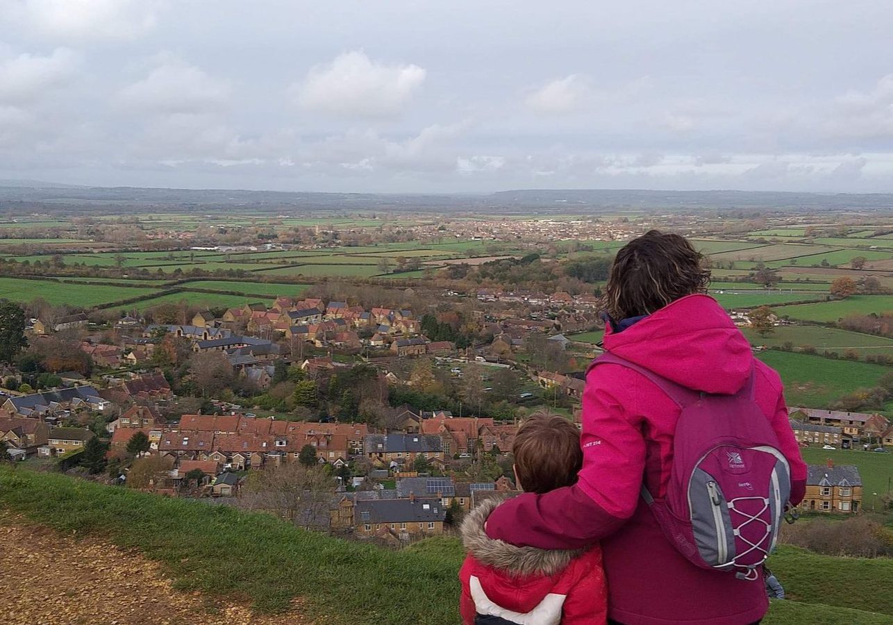 A woman in a pink coat with a small pink backpack on her back. Her left arm is held around a small boy in a red, white and navy colour block coat. They are looking out over the countryside. The sky is grey with low white clouds and small villages are dotted in amongst green fields.