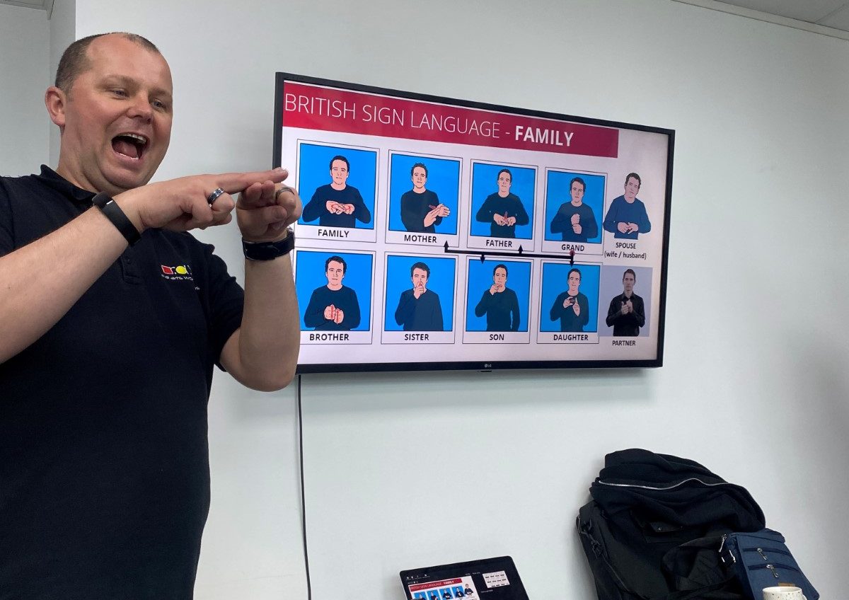 A man is stood next to a TV screen with images of BSL signs on it. The man is using his hands to show how to recreate the signs.