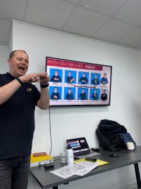 A man is stood next to a TV screen with images of BSL signs on it. The man is using his hands to show how to recreate the signs.