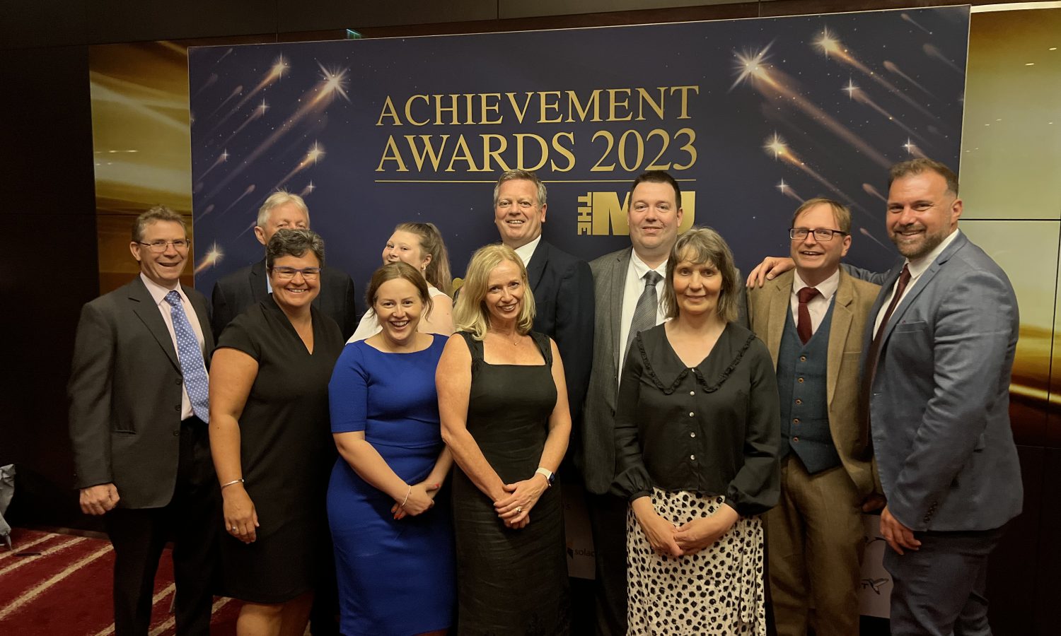 Eleven people stood smiling in front of an awards banner which reads "achievement awards 2023". They are smartly dressed