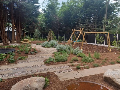 An image looking down the garden. Wooden supports are at the far end holding swings. Slabs make a pathway and shrubs are growing all around.