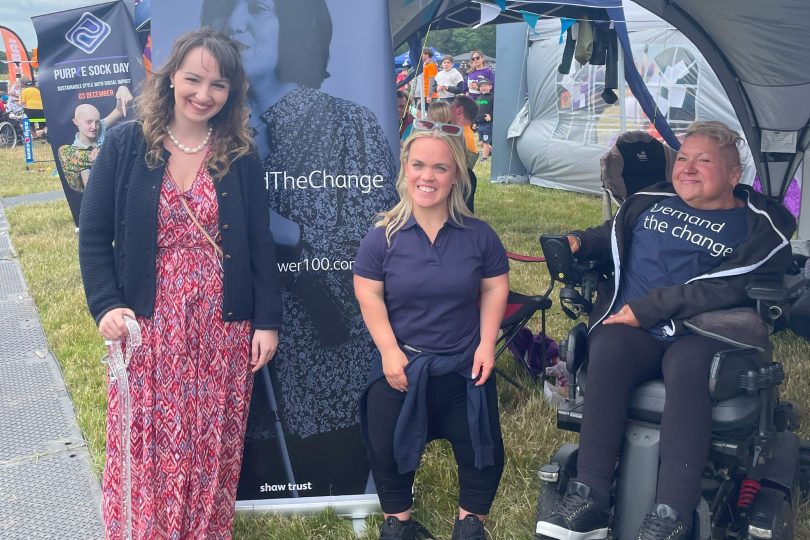Beth Kume Holland, Ellie Simmonds and Clare Gray smile at the camera at the Shaw Trust stand, in front of a Demand the Change pull up banner.