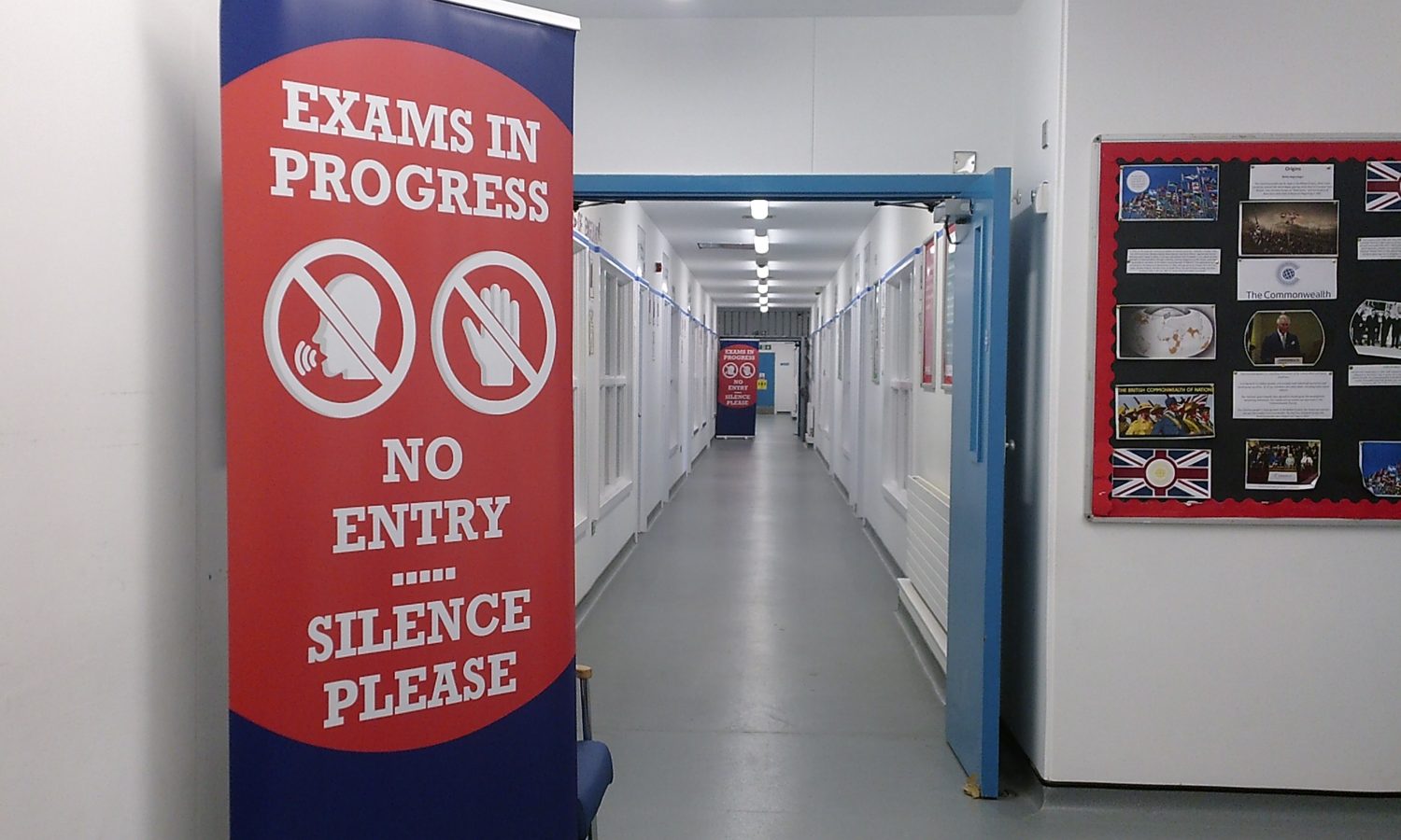 The image looks down a long, clean white corridor. A large pull up banner is in the foreground which says "Exams in Progress. No entry, silence please."