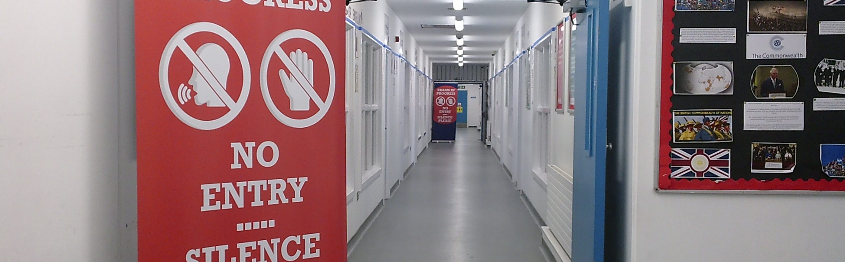 The image looks down a long, clean white corridor. A large pull up banner is in the foreground which says "Exams in Progress. No entry, silence please."