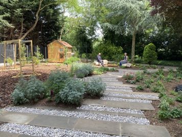 An image looking down the garden. Wooden supports are at the far end holding swings. Slabs make a pathway and shrubs are growing all around.