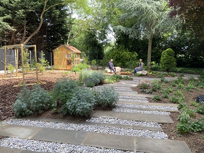 An image looking down the garden. Wooden supports are at the far end holding swings. Slabs make a pathway and shrubs are growing all around.