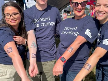 Four members of the Foundation team holding their arms into the middle. Each are wearing a navy blue tshirt and blue tattoo reading Demand The Change