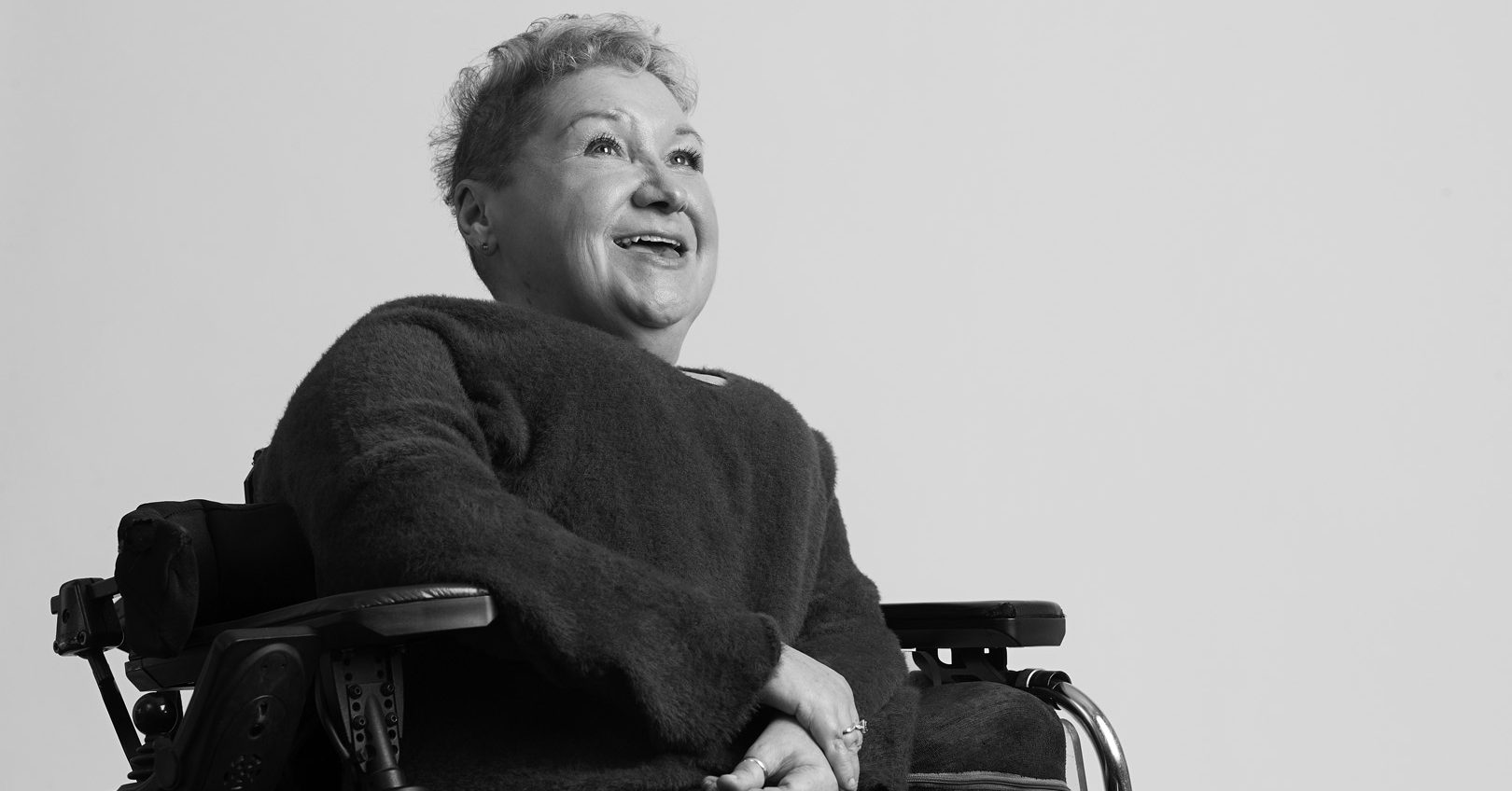 A black and white image of Clare Gray. A woman with short hair and a big smile sat in her wheelchair.